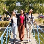 Full body of cheerful multiracial female friends strolling together on wooden footbridge over river in city on sunny summer day