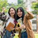 Two excited young Asian female college students are raising their fists in celebration, smiling happily while standing outdoors on a sunny day. university life and friendship concepts