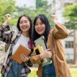 Two excited young Asian female college students are raising their fists in celebration, smiling happily while standing outdoors on a sunny day. university life and friendship concepts